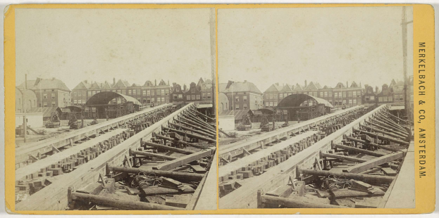 Schwarz-weiß-Foto einer Zugstrecke mit Gebäuden im Hintergrund, Text 'Merkelbach & Co, Co. Amsterdam' rechts und verstreute hölzerne Gegenstände vorn.
