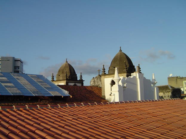 Eine Stadtansicht mit mehreren Gebäuden im Vordergrund unter einem blauen Himmel, die Solarpanels auf einem Dach zeigt.