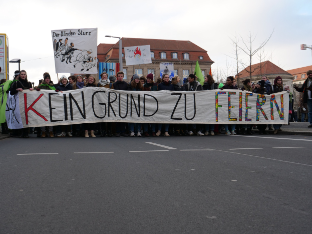 Gruppe von Menschen auf einer Straße mit einem Banner 'Kein Grund zu Feiern' im Protest, mit Gebäuden, Bäumen und einem klaren Himmel im Hintergrund.