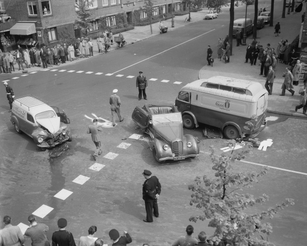 Schwarz-weißes Bild eines Autounfalls an einer Straßenkreuzung mit mehreren Fahrzeugen, darunter ein Lieferwagen, und einer Gruppe von Schaulustigen; Gebäude, Bäume, Strommasten und Schilder sind im Hintergrund zu sehen.
