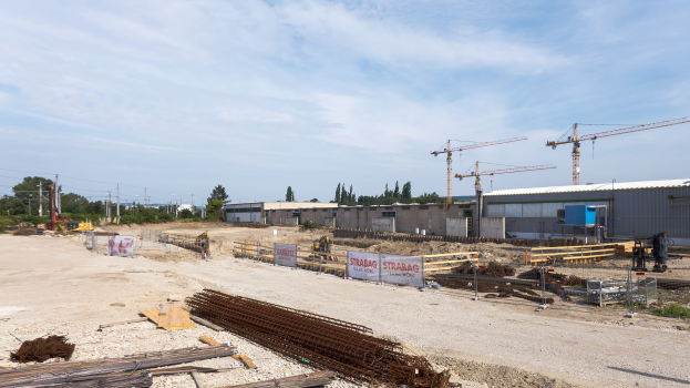 Baustelle mit verschiedenen Materialien und Geräten, darunter Eisenstangen, Holzplanken, Pfähle, Drähte, Kräne und Gebäude unter einem bewölkten Himmel.