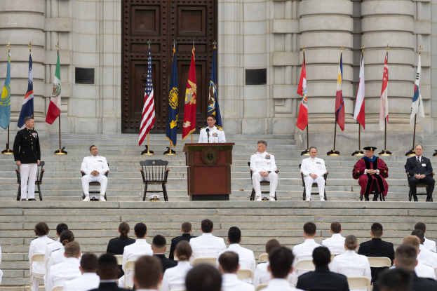 Gruppe von Menschen in weißen Uniformen auf den Stufen eines Gebäudes mit einem Podium, das von Fahnen flankiert ist, vor einem großen Gebäude mit einer Tür, das eine Ehrenzeremonie vermuten lässt.