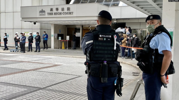 Zwei Polizisten in Uniform und Masken vor einem Gebäude am Hong Kong International Airport, umgeben von Menschen mit Kameras, mit Text auf dem Gebäude und gespannten Bändern im Hintergrund.