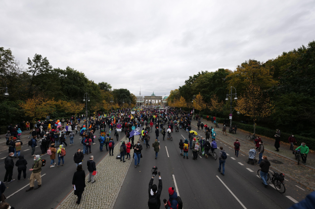 Eine große Gruppe von Menschen marschiert eine baumbestandene Straße entlang, mit Laternenmasten, und hält Kameras, mit einem Gebäude und einem klaren Himmel im Hintergrund.