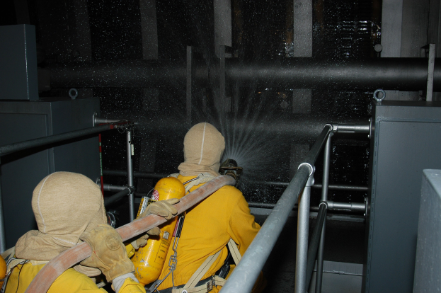 Zwei Feuerwehrleute in gelben Uniformen, Helmen und Handschuhen spritzen Wasser aus einem Schlauch auf ein Metallgeländer mit Metallstangen und anderen Gegenständen im Hintergrund.