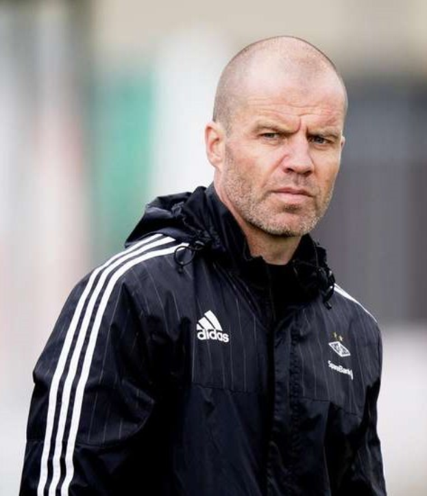 Glatzköpfiger Mann in einem blauen Jackett mit weißen Streifen und weißem Kragen, der auf einem Fußballfeld mit gefalteten Händen und ernster Miene steht, unscharfer Hintergrund.