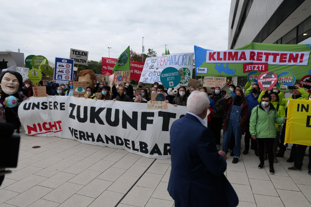 Eine Gruppe von Menschen protestiert gegen den Klimawandel in Deutschland, sie halten Schilder und Banner vor einem Gebäude, während ein Fotograf das Ereignis dokumentiert, vor einem Hintergrund aus Bäumen, Pfählen und einem klaren blauen Himmel.