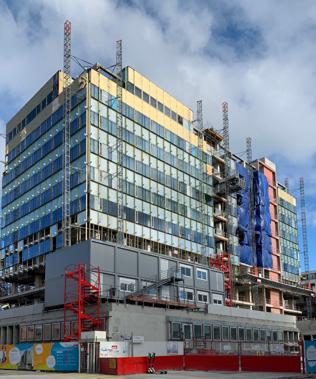Großes Gebäude im Bau in einer Stadt mit Glasfenstern, Metallrahmen, einer Treppe, einem Zaun, einer Tafel, Pfosten, einem Straßenpfosten, Bäumen und einem bewölkten Himmel.