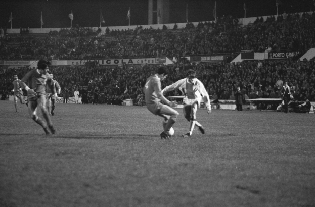 Schwarzes und weißes Bild von Männern, die auf einem Feld in einem Stadion Fußball spielen, mit Zuschauern auf den Tribünen und Fahnen im Hintergrund.