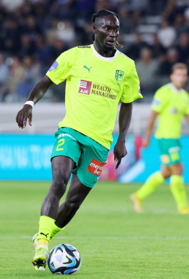 Ein Fußballer in gelbem T-Shirt und grünen Shorts ist im Einsatz auf dem Feld, mit einer blauen Tafel und unscharfen Figuren im Hintergrund.