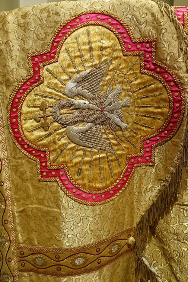 Nahaufnahme eines Priestergewandes mit aufwendiger Gold- und Rotstickerei vor einem neutralen Hintergrund.