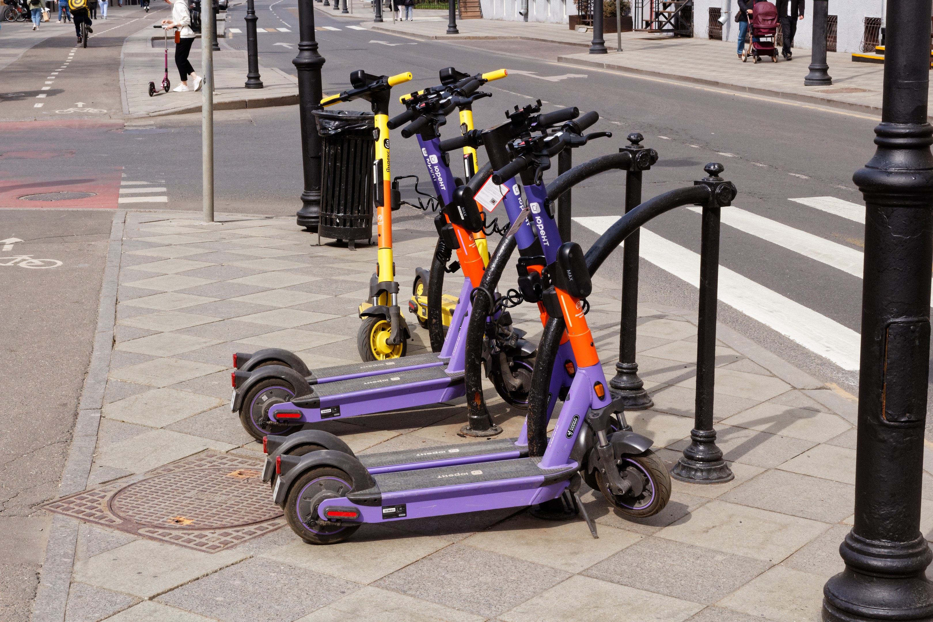 Eine Reihe von Elektro-Scootern, die entlang einer Straße geparkt sind, mit Fußgängern, Radfahrern, einem Müllcontainer im Vordergrund und Gebäuden, Bäumen und anderen Objekten im Hintergrund.