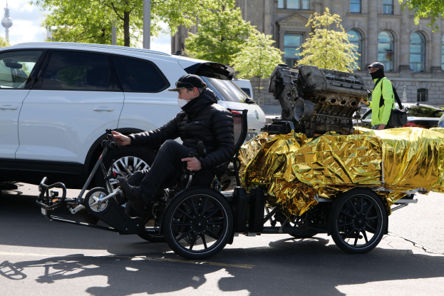 Ein Mann im Rollstuhl mit einem großen Motor auf dem Rücken, umgeben von Fahrzeugen auf einer Straße mit Bäumen, Gebäuden, Masten und einem klaren blauen Himmel im Hintergrund; er trägt eine schwarze Jacke, eine Mütze und hält ein Objekt in der Hand.