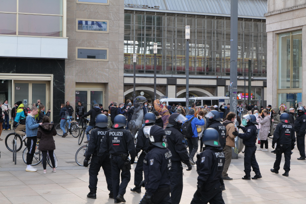 Eine Gruppe von Polizisten in Helmen steht vor einer Menge, die Fahrräder hält, mit Glaswandgebäuden, Laternenmasten und Fahrzeugen im Hintergrund.