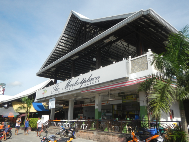 Marktplatz Öffentlicher Markt in Koh Samui, Thailand mit Menschen, geparkten Fahrzeugen, einem Metallzaun, Bäumen, einer Plane, Masten, einem Dach mit Deckenleuchten und einem bewölktem Himmel.