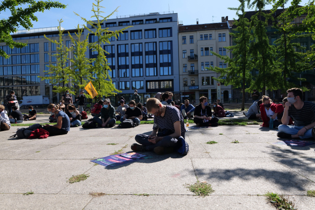 Gruppe von Menschen, die vor einem Gebäude auf dem Boden sitzen, während einer Demonstration in Berlin, einige tragen Masken, mit verstreuten Taschen und Gegenständen, umgeben von Bäumen unter einem klaren blauen Himmel.