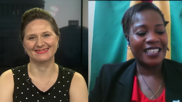 Zwei Frauen, die während eines Videoanrufs lächeln, eine in einem schwarzen und weißen Kleid und die andere in einer unbestimmten Farbe, mit einer Fahne, die an der Wand hinter ihnen in einer öffentlichen Einrichtung hängt.