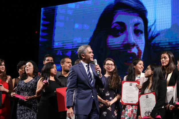 Ein Mann im Anzug und mit Krawatte spricht auf einer Bühne, während das Publikum Zertifikate hält und im Hintergrund ein Bild einer Frau auf einem Bildschirm zu sehen ist.