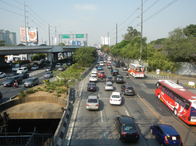 Eine belebte städtische Straße mit Autos, Bussen, Strommasten, Bäumen, Gebäuden, Werbetafeln und einer Brücke unter einem bewölkten Himmel.