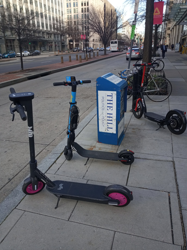 Zwei Elektro-Scooter neben einer Stadtstraße mit Fahrrädern, einem Mülleimer und anderen städtischen Elementen im Vorder- und Hintergrund.