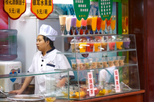 Ein Koch steht neben einem Tisch mit Saftgläsern, Schalen mit Süßigkeiten und Bannern darauf, mit einem Wasserspender und Kühlschrank im Hintergrund.