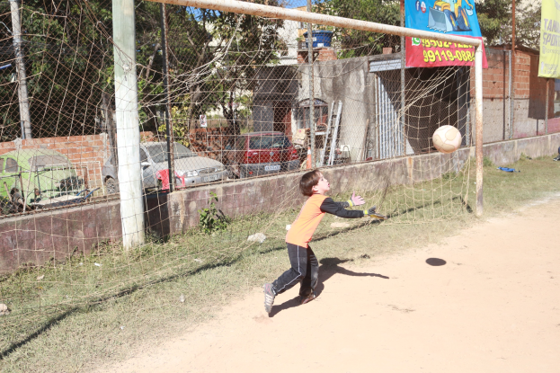 Ein junger Junge kickt einen Füüball in ein Netz, mit Bannern, Gebäuden, Bäumen, Fahrzeugen und einem klaren blauen Himmel im Hintergrund.
