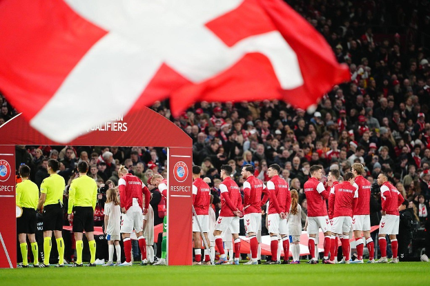 Gruppe von Menschen auf einem Fußballfeld mit einer roten und weißen Flagge im Vordergrund, einem Bogen mit der Aufschrift "Bayern Munich vs. Bayern Munich Wetten & Vorschau" im Hintergrund und einer großen Menge im Stadion.