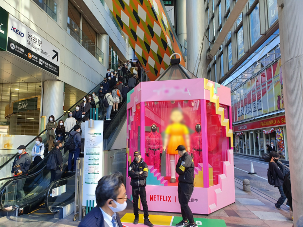 Eine Gruppe von Menschen steht vor einem Gebäude mit Glaswänden, Säulen und Texttafeln, einige tragen Masken, während eine Rolltreppe mit Menschen zu sehen ist. In der Mitte steht eine pinkfarbene Box mit Schaufensterpuppen und der Aufschrift "Netflix' neue Netflix-Bühne in Tokio."