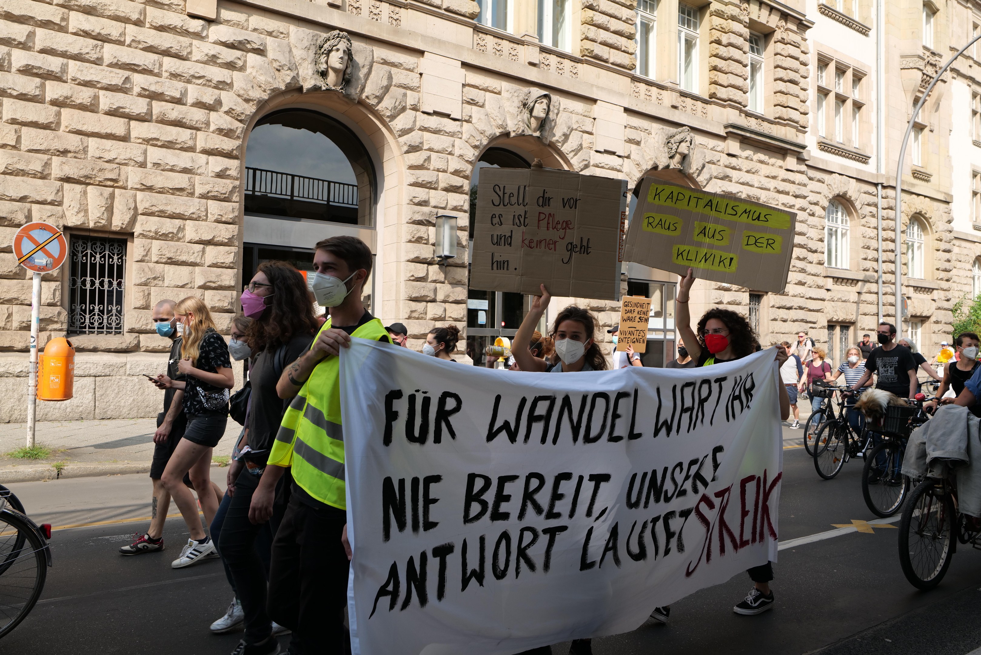Eine Gruppe von Demonstranten marschiert durch eine Straße in Berlin, einige halten Schilder und andere fahren Fahrräder, im Hintergrund ein historisches Gebäude mit Bögen, Säulen und Skulpturen.
