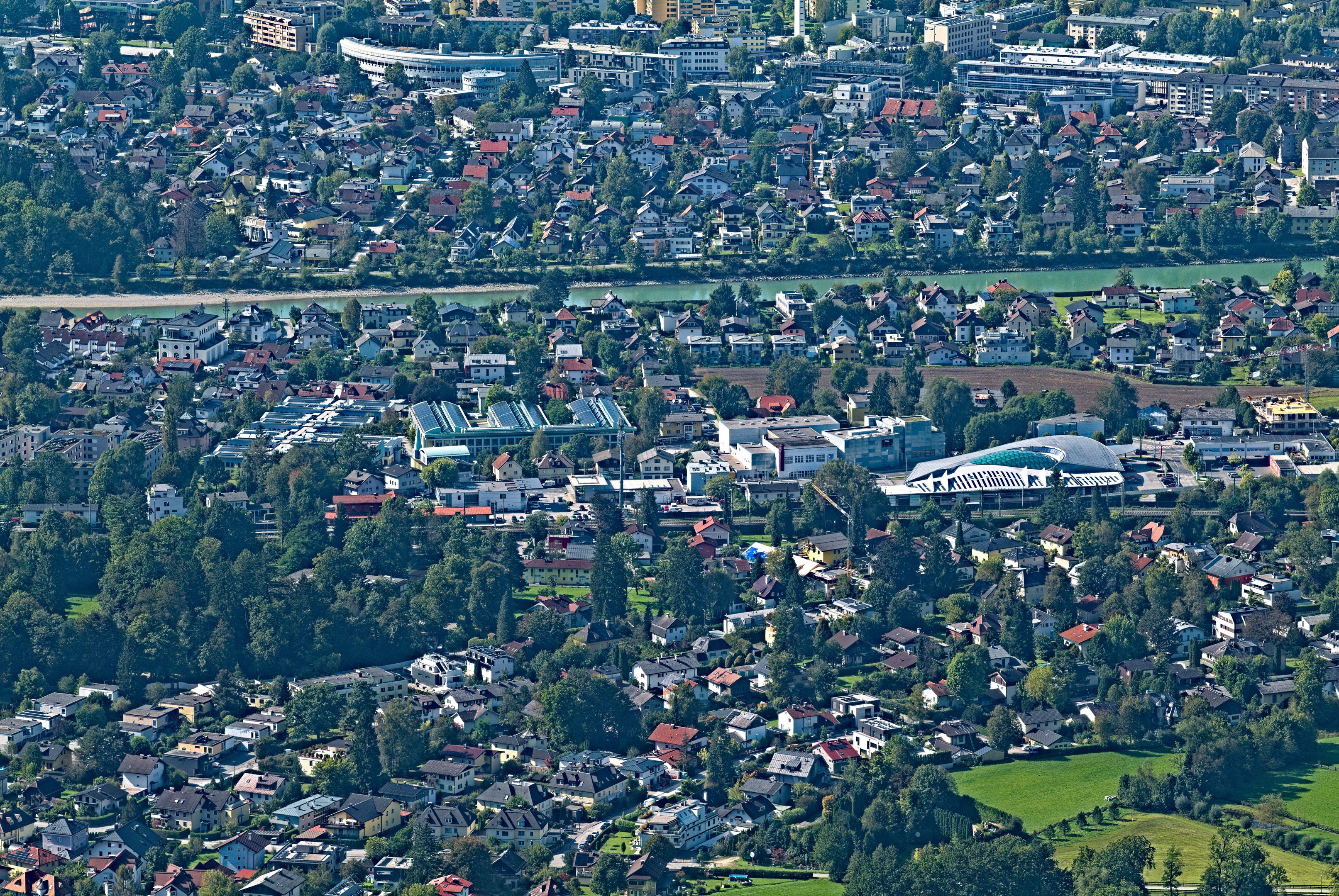 Luftaufnahme einer Stadt mit dichtem Baumbestand, Grünflächen, Straßen, Fahrzeugen und Wasser, umgeben von einem großen Gebäude mit Gras und Bäumen.