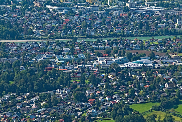 Luftaufnahme einer Stadt mit dichtem Baumbestand, Grünflächen, Straßen, Fahrzeugen und Wasser, umgeben von einem großen Gebäude mit Gras und Bäumen.