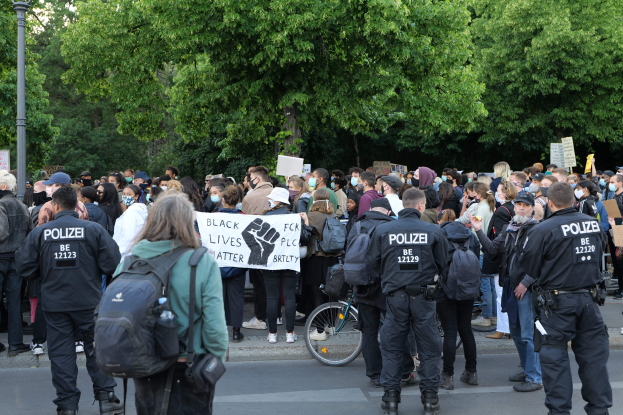 Eine große Gruppe von Menschen, die auf der Straße stehen und Schilder halten, einige tragen Mützen und Taschen, vorne ein Fahrrad und im Hintergrund Bäume und ein Mast.