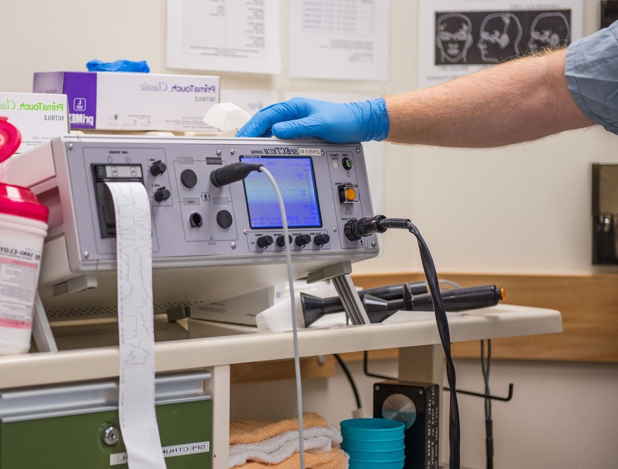 Eine Person in blauen Handschuhen arbeitet an einem medizinischen Gerät auf einem Tisch in einem unordentlichen Krankenhauszimmer mit Kisten, Tassen und Papieren an den Wänden.