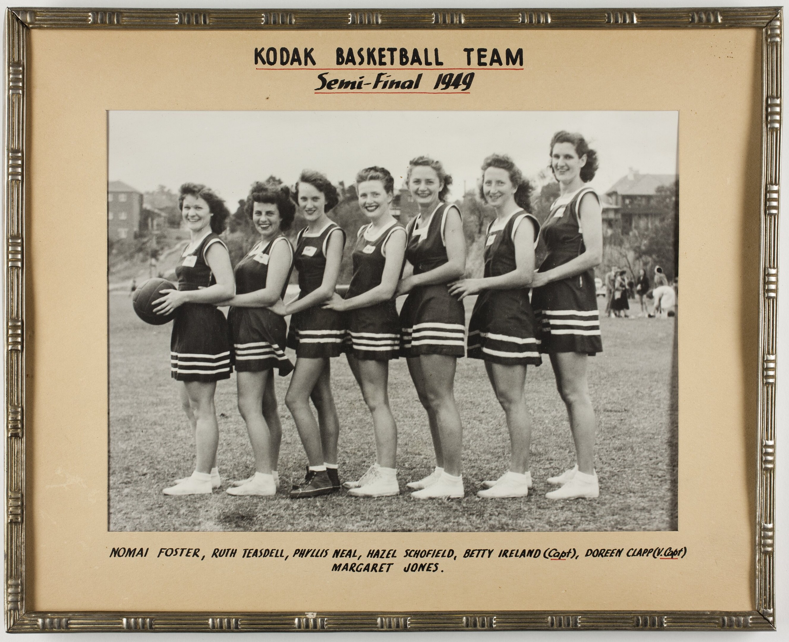 Schwarzes und weißes Foto einer Gruppe von Frauen, die strahlend einen Basketball halten, mit Menschen, Bäumen, Gebäuden und Himmel im Hintergrund, beschriftet mit "Kodak Basketball Team Semi-Final 1949".