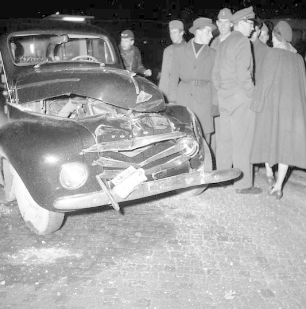 Schwarz-Weiß-Bild Eines Schwer Beschädigten Autos Am Straßenrand Mit Einem Zerdrückten Kühlergrill Und Zerbrochenen Fenstern, Umgeben Von Menschen, Die Die Szenerie Begutachten.