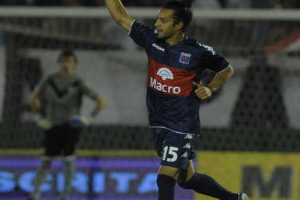 Ein Fußballspieler feiert nach einem Tor auf dem Boden, mit jubelnden Zuschauern und einer Person im Hintergrund und einer Tafel mit Text dahinter, alles mit einem unscharfen, energetischen Hintergrund.