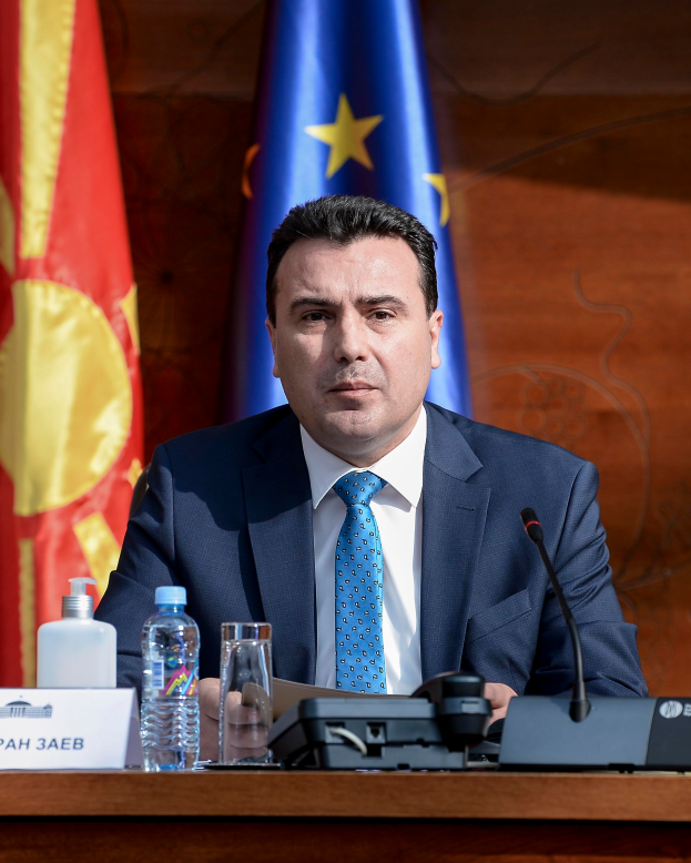 Ein Mann im Anzug und mit Krawatte sitzt an einem Tisch mit Mikrofon, Flaschen, einem Glas und einer Namensschilder, mit Flaggen im Hintergrund.