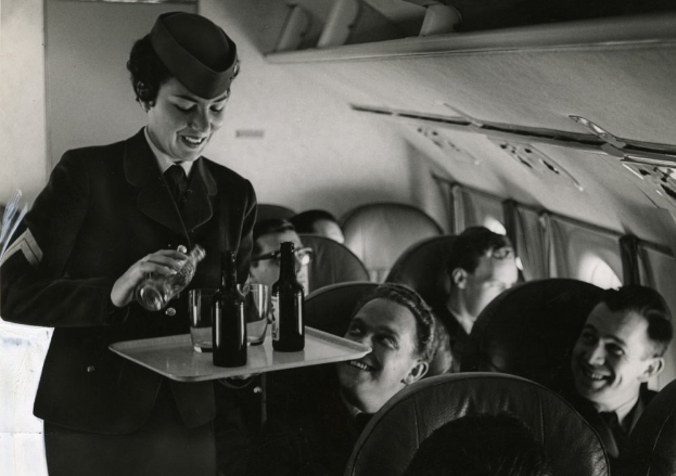 Schwarzes und weißes Bild einer Flugbegleiterin, die Getränke von einem Tablett an sitzende Passagiere auf einem Flugzeug serviert.