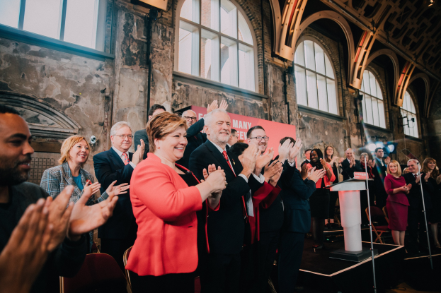 Eine Gruppe von Menschen, die vor einem Publikum klatschen, mit einem Podium, einem Mikrofon, Stühlen, einer Wand, einem Banner mit der Aufschrift "Covid-19 New York City Mayor Bill de Blasio" und Fenstern im Hintergrund.