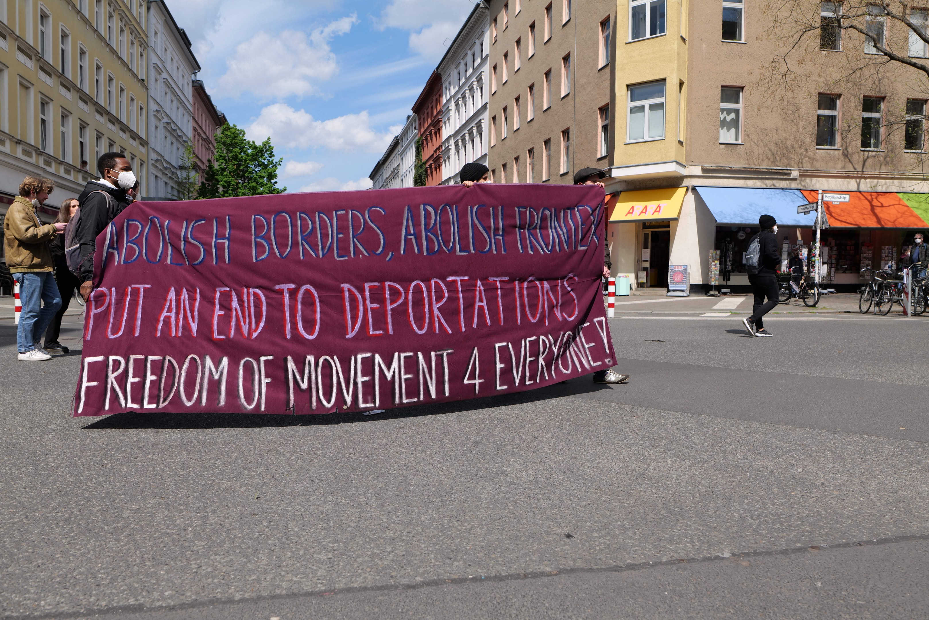 Eine Gruppe von Menschen marschiert mit einem Banner, auf dem "Abolish Borders, Abolish Frontiers, Put an End to Deportations, Freedom of Movement 4 Everyone" steht, eine Straße entlang. Im Hintergrund sind Gebäude, Bäume, Fahrräder und ein bewölkter Himmel zu sehen.