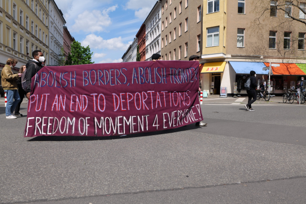Eine Gruppe von Menschen marschiert mit einem Banner, auf dem "Abolish Borders, Abolish Frontiers, Put an End to Deportations, Freedom of Movement 4 Everyone" steht, eine Straße entlang. Im Hintergrund sind Gebäude, Bäume, Fahrräder und ein bewölkter Himmel zu sehen.