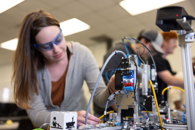 Eine Frau mit Schutzbrille arbeitet an einem Roboter in einem Labor, umgeben von anderen, mit Werkzeugen und Komponenten auf dem Tisch unter Deckenbeleuchtung.