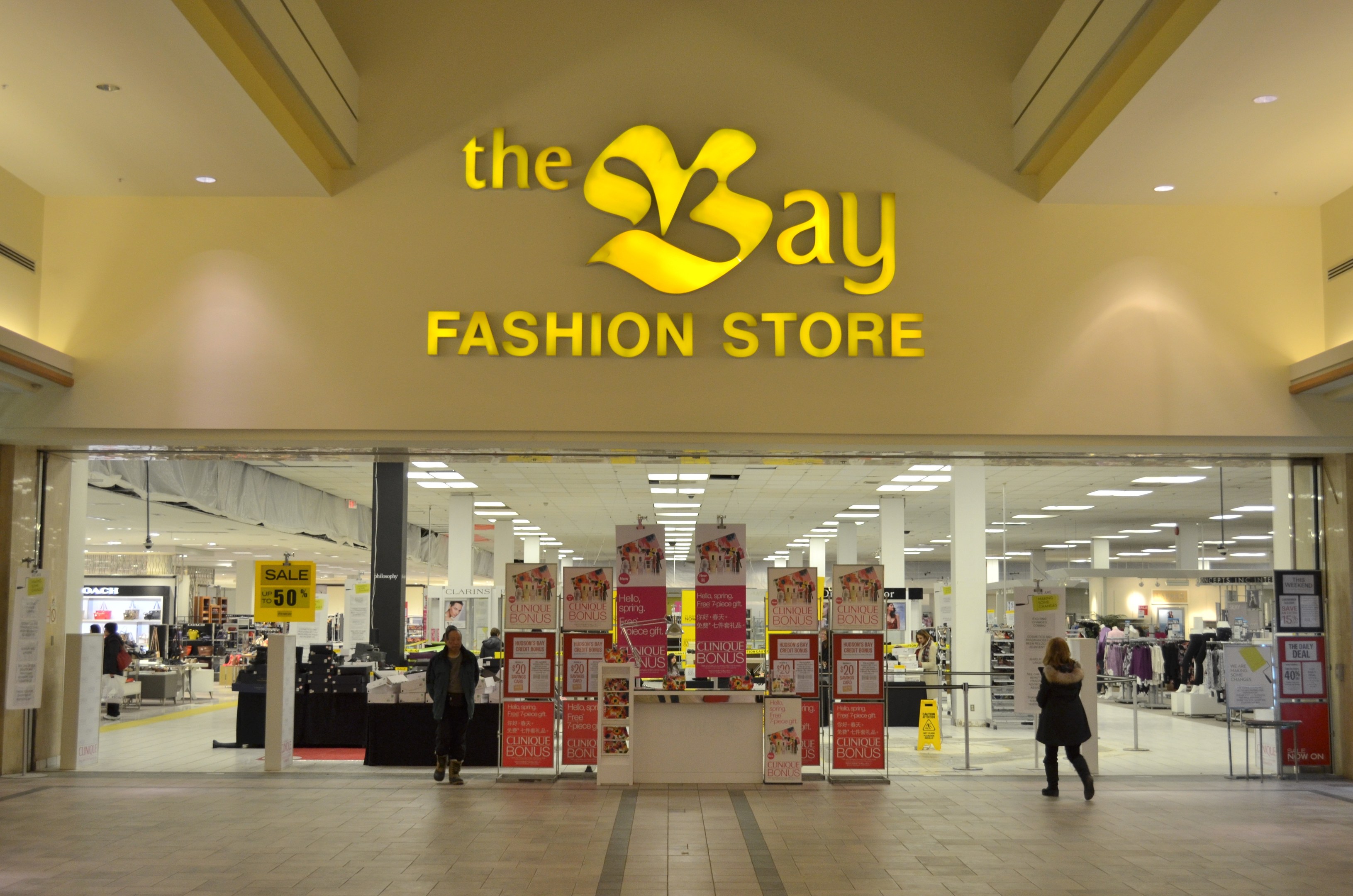 Außenansicht eines Bay Fashion Stores in einem Einkaufszentrum mit Passanten, Wandtexttafeln, Säulen, Deckenleuchten und Hintergrundauslagen.