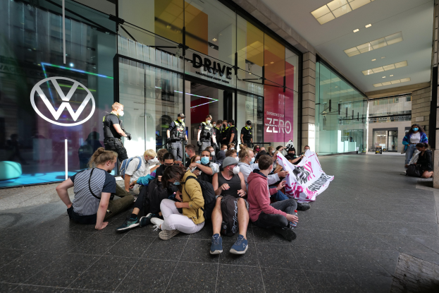 Gruppe von Menschen, die vor einer Volkswagen-Niederlassung auf dem Boden sitzen, einige halten ein Banner, mit Glaswand-Gebäude und Deckenbeleuchtung.