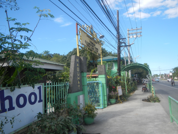 Einfahrt zu einer Schule mit einem "Schule"-Schild, Fahrzeuge auf der Straße, ein Torbogen, ein Tor, ein Namensschild, Pflanzen, Bäume, Strommasten mit Drähten und eine bewölkte Himmel im Hintergrund.