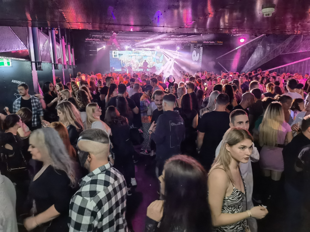 Eine große Menge steht in einem Nachtclub mit Deckenlichtern und Säulen, mit einem Bildschirm im Hintergrund, der als Absolut Nightlife-Event gekennzeichnet ist.
