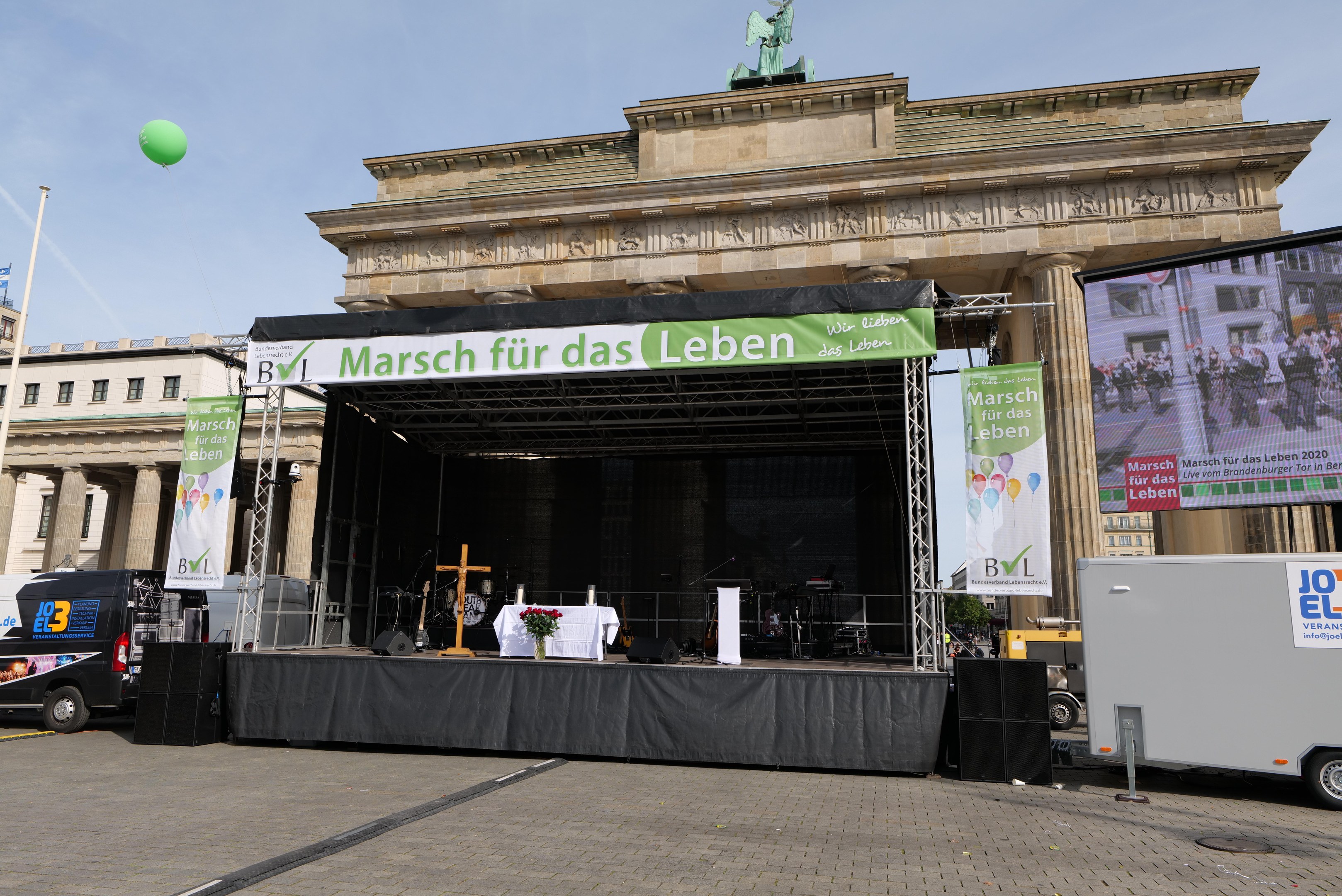 Bühne vor dem Brandenburger Tor in Berlin, Deutschland, mit einem Tisch mit einer Blumenvase, einem Kreuzsymbol, Transparenten mit Text, Lautsprechern und anderen Gegenständen sowie Fahrzeugen auf der Straße und Gebäuden im Hintergrund.
