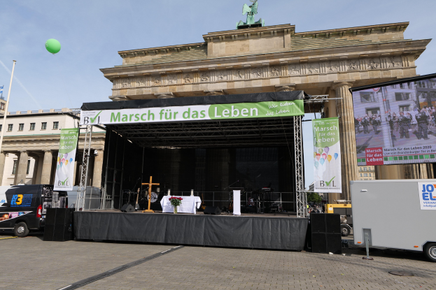 Bühne vor dem Brandenburger Tor in Berlin, Deutschland, mit einem Tisch mit einer Blumenvase, einem Kreuzsymbol, Transparenten mit Text, Lautsprechern und anderen Gegenständen sowie Fahrzeugen auf der Straße und Gebäuden im Hintergrund.