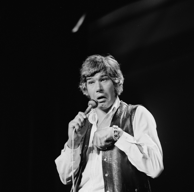 Ein Mann in weißem Hemd und schwarzer Weste singt leidenschaftlich in ein Mikrofon, seine Augen geschlossen und das Haar im klassischen Rocker-Look gestylt.