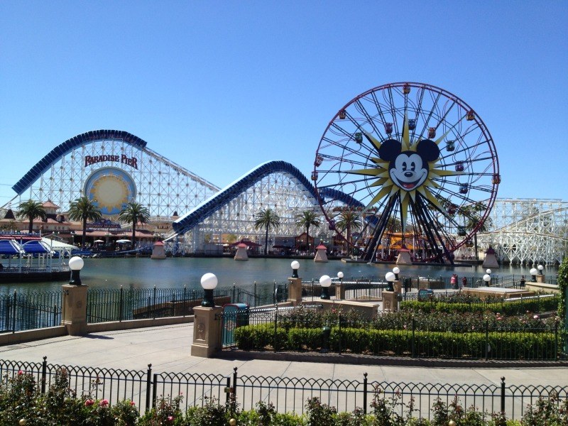 Luftaufnahme von Disney California Adventure mit seinem Riesenrad und Achterbahn, umgeben von Grünflächen und Laternenmasten, mit Wasser im Vordergrund und einem klaren blauen Himmel.